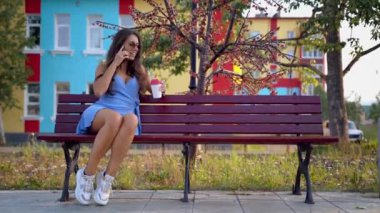 Güneş gözlüklü, mavi elbiseli, bankta oturan güzel bir esmer. Kahve içer ve telefonda konuşur..