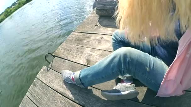Menina loira em jeans azul senta-se na borda de um velho cais de madeira — Vídeo de Stock