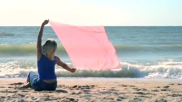 Chica con una bufanda rosa está sentado en la orilla de arena de la playa — Vídeo de stock