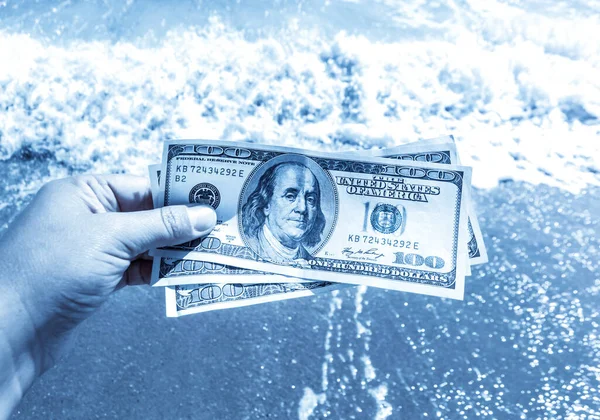 Girl holding money bill of 300 dollars on background of sea ocean waves with white foam and sand wet beach close-up. Hand wave sea ocean money dollars vacation. Concept finance money holiday traveling