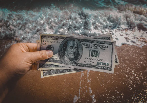 Girl holding money bill of 300 dollars on background of sea ocean waves with white foam and sand wet beach close-up. Hand wave sea ocean money dollars vacation. Concept finance money holiday traveling