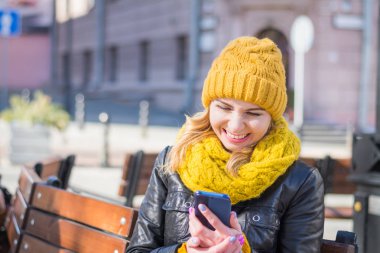 bir örme şapka ve eşarp güzel kadın bir tezgah üzerinde oturur ve onun Smartphone ekranına bakarken gülüyor