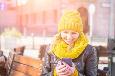 bir örme şapka ve eşarp güzel kadın bir tezgah üzerinde oturur ve onun Smartphone ekranına bakarken gülüyor