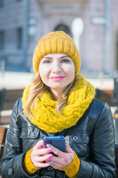 portrait of a young attractive woman in a knitted hat and knitted scarf, which sits on a bench in the city park