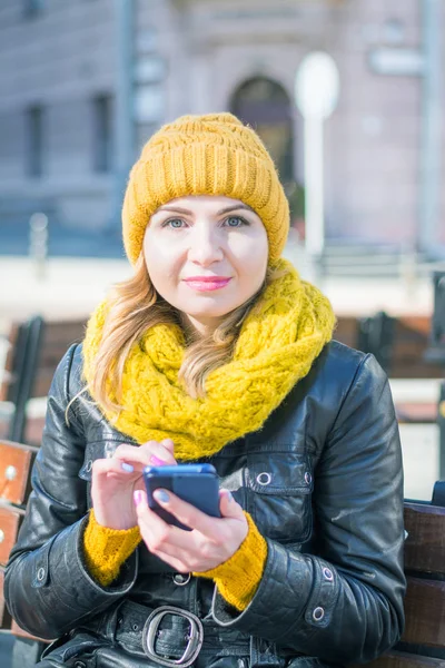 Portrait of a young attractive woman in a knitted hat and knitted scarf, which sits on a bench in the city park — Foto de Stock