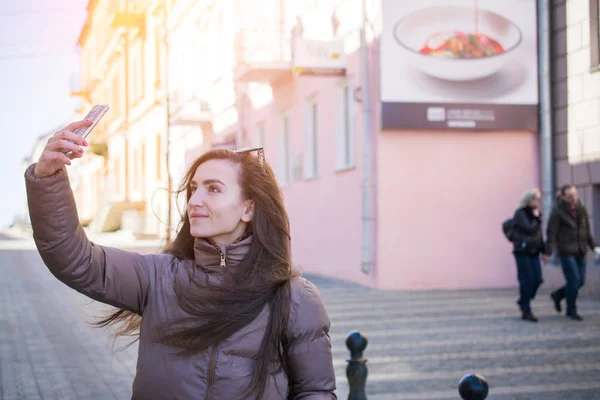 Outdoor street style image of young trendy woman making selfie on the city street