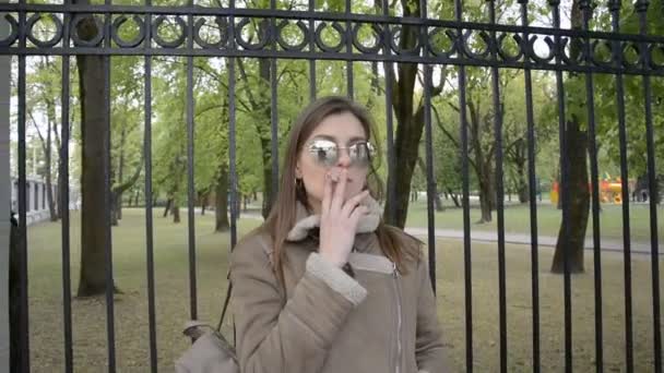 Beautiful girl in a jacket and sunglasses smokes in the park — Stock Video