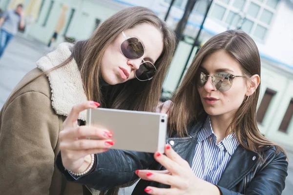 Twee stijlvolle meiden nemen een selfie op hun smartphone in een stads straat en glimlachen naar de camera — Stockfoto