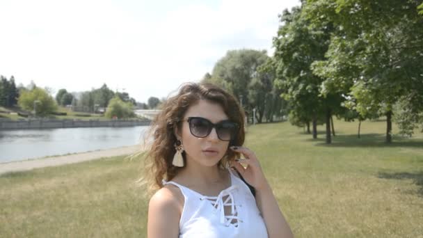 Menina bonita com cabelo encaracolado olhando para a câmera em um parque da cidade e com um rio no fundo — Vídeo de Stock