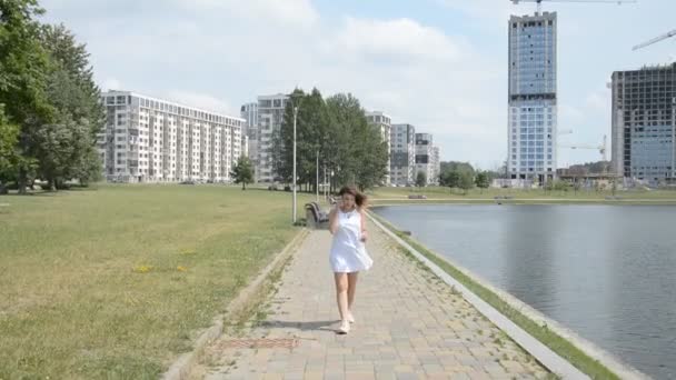 Jovem atraente em um vestido de verão branco caminha pelo parque e fala sobre o telefone — Vídeo de Stock