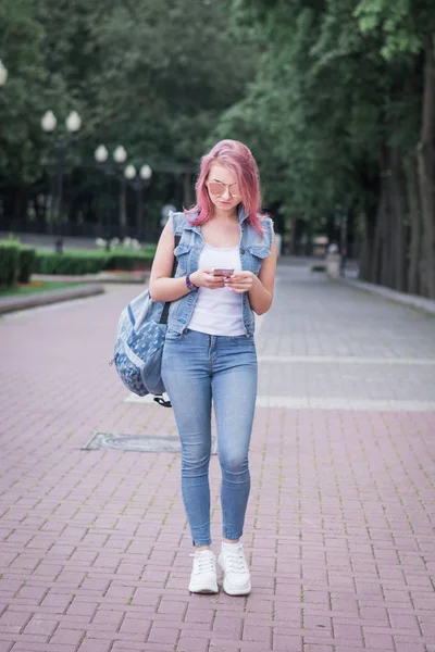 Mujer urbana joven con peinado moderno usando smartphone caminando en la calle en un parque urbano — Foto de Stock