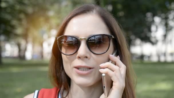 Mooi jong meisje in zonnebril praten met haar vriend op een mobiele telefoon in een zomerpark — Stockvideo