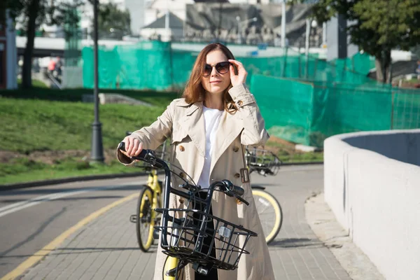 Stylish girl student in a raincoat and sunglasses riding a city on a rented bike — Stock Photo, Image