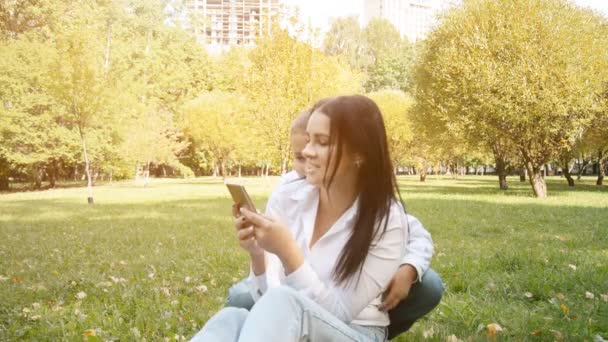 Современная концепция семейной жизни: красивая мать и ее восьмилетний сын сидят в парке на газоне и используют телефон для общения в Интернете — стоковое видео