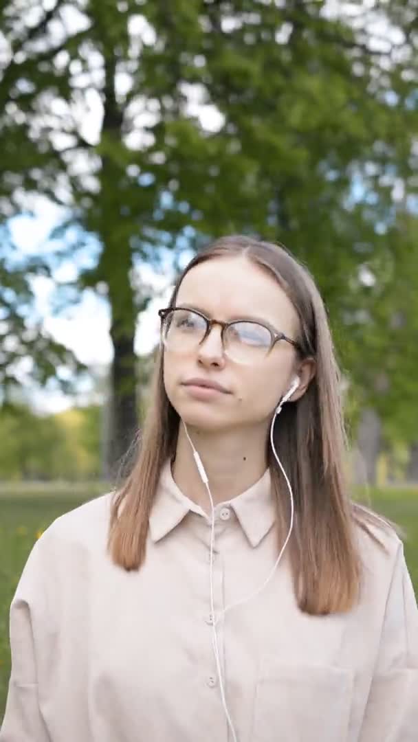 Bella ragazza studente in occhiali e cuffie bianche sulla natura ascolta la musica — Video Stock