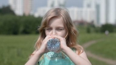 Küçük güzel bir kızın portresi doğada kızaran bir yaz gününde plastik bir şişeden su içer.