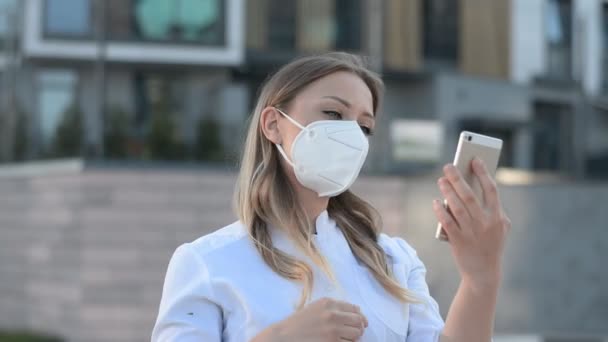 Νεαρή γιατρός κορίτσι σε μια προστατευτική μάσκα κοιτάζει το τηλέφωνο και ισιώνει τα μαλλιά στο δρόμο της πόλης του — Αρχείο Βίντεο