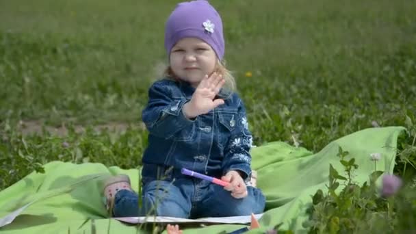 Bambina seduta in un parco sull'erba verde che guarda la macchina fotografica e saluta — Video Stock