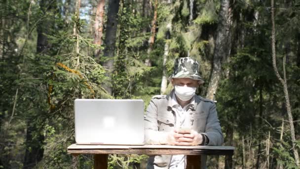 Coronovirus pandemie in de wereld, een man zit in een bos met een beschermend masker en praat aan de telefoon, een laptop staat in de buurt, een werkplek tijdens de epidemie — Stockvideo