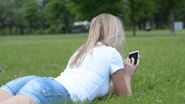 Χαριτωμένο γυναίκα διαβάζει το μήνυμα κειμένου στο κινητό τηλέφωνο στο γρασίδι πάρκο πίσω άποψη — Αρχείο Βίντεο