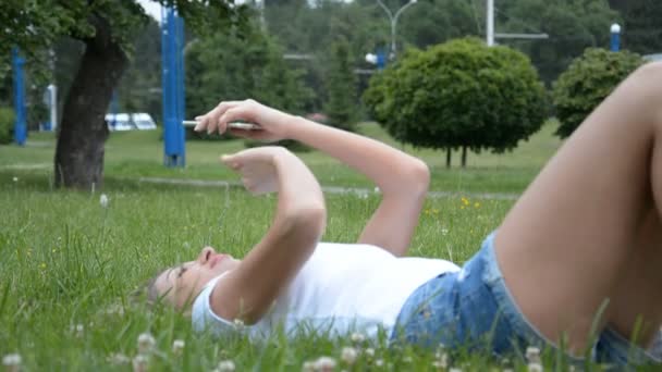 Egy lány okos telefont használ a park füvén. — Stock videók