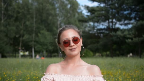 Кавказька дівчинка в сонцезахисних окулярах дивиться на камеру і посміхається в парку в літній день — стокове відео