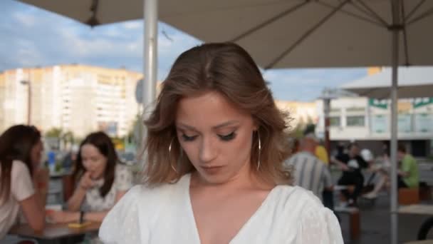 Retrato de una hermosa chica en un vestido blanco y pendientes grandes en un café de verano — Vídeos de Stock