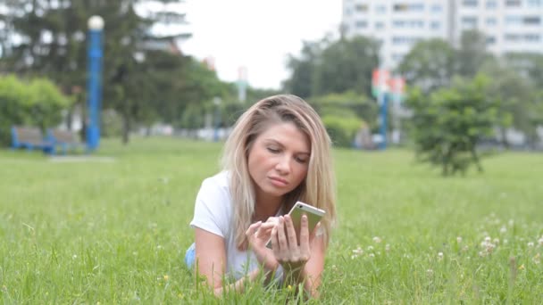 Mooie blonde vrouw ligt op een gazon en schrijft een bericht per telefoon — Stockvideo