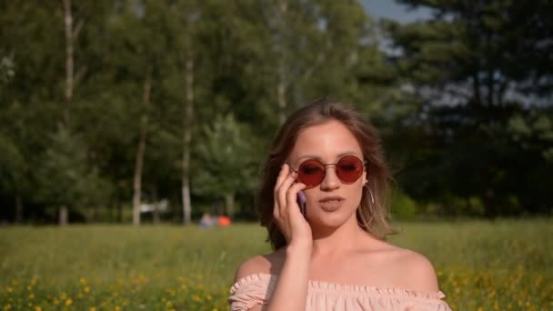 Портрет красивої стильної дівчини в літньому одязі, яка сидить в парку на газоні і розмовляє по телефону, вільний простір для тексту — стокове відео