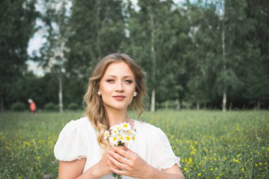 Beyaz elbiseli ve elinde papatya demeti olan büyük beyaz şapkalı güzel bir kız yaz parkındaki fotoğrafçıya poz veriyor.