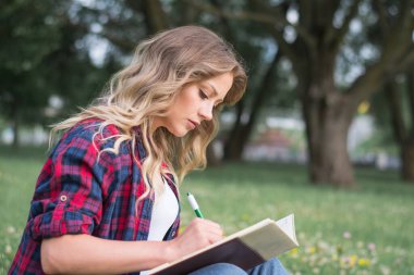 Güzel, mutlu genç öğrenci kız sırt çantasının yanındaki yeşil çimenlerde oturuyor, yaz günü kampüsün yakınındaki ağacın altında çanta.