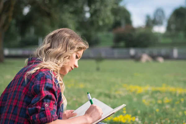 Güzel, mutlu genç öğrenci kız sırt çantasının yanındaki yeşil çimenlerde oturuyor, yaz günü kampüsün yakınındaki ağacın altında çanta.