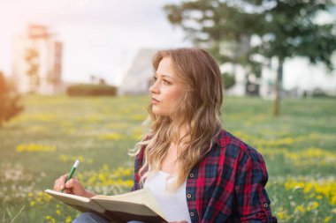 Genç beyaz kız bir yaz parkında kampüs çimlerinde otururken deftere notlar yazıyor.