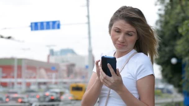 Hübsche junge kaukasische Frau, die auf dem Handy SMS schreibt, während sie darauf wartet, am Straßenrand abgeholt zu werden — Stockvideo