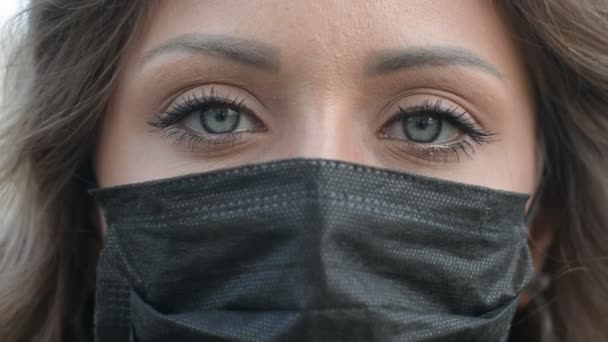Closeup olhos tristes de jovem mulher caucasiana em uma máscara médica preta piscando e olhando para a câmera — Vídeo de Stock
