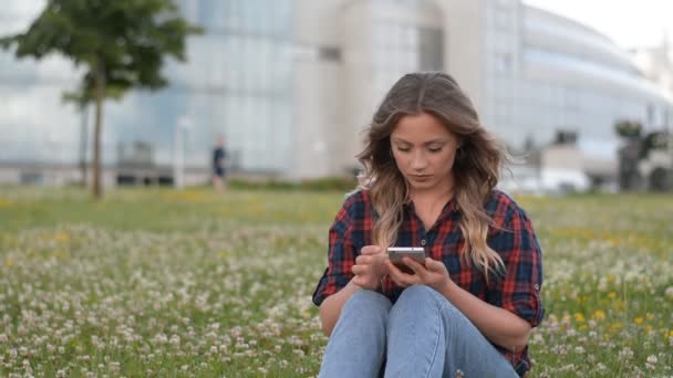 Joven hermosa caucásica chica sentada en el césped en el parque usando el teléfono — Vídeo de stock