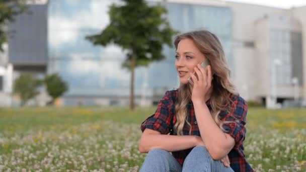 Menina hipster bonita jovem senta-se à noite na grama em um parque da cidade no fundo de um edifício moderno e falando ao telefone — Vídeo de Stock