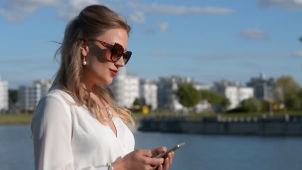 Güneş gözlüklü ve beyaz elbiseli zarif ve güzel bir kadın nehrin kenarında duruyor ve arkadaşlarına bir mesaj yazıyor. — Stok video
