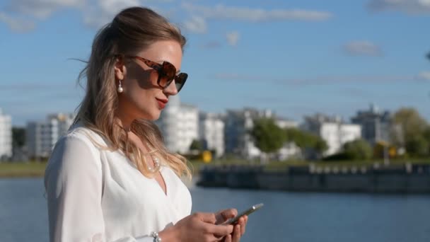 Jovem elegante e bonito em óculos de sol e um vestido branco fica perto do rio e escreve uma mensagem em um smartphone para seus amigos — Vídeo de Stock