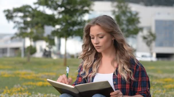 Hermosa niña caucásica sentada en la hierba en el parque y escribiendo en el diario — Vídeo de stock