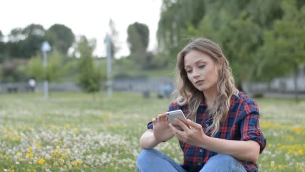 Nahaufnahme Portrait von jungen schönen Mädchen sitzt auf Rasen im Park mit dem Handy — Stockvideo