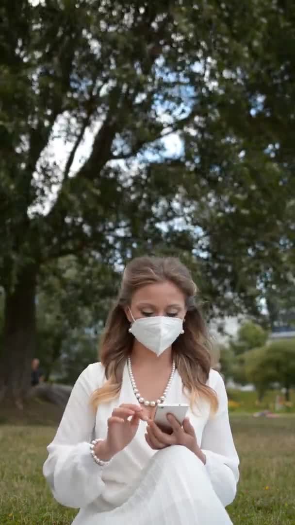 Elegante stijlvolle vrouw in een witte jurk en een beschermend masker op haar gezicht zit op het gazon in het park en kijkt in haar smartphone — Stockvideo