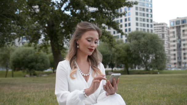 Menina bonita em um vestido de noite branco vestido em estilo retro senta-se no gramado e escreve uma mensagem em seu telefone ou lê as últimas notícias — Vídeo de Stock