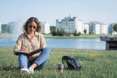 Beyaz kız sınavlara hazırlanıyor çimenlerde oturuyor ve not defterine notlar yazıyor.