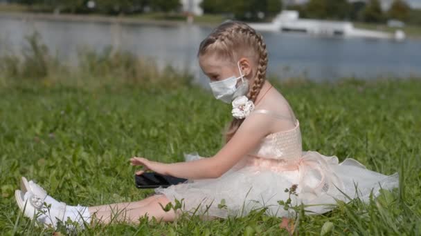 Kleines Mädchen mit Schutzmaske und Handy in der Hand sitzt auf dem Rasen — Stockvideo