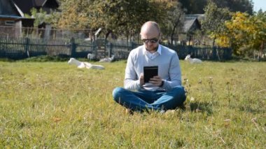 İşletme müdürü köyde çimlerin üzerinde oturuyor ve keçilerin arka planına karşı tabletine bir şeyler yazıyor.