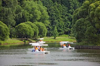 Moscow / Russia  07 16 2019: Tsaritsino Upper Pond with pedal boats tourists Catamarans near Round Island on green trees background in Tsaritsyno Park Museum on summer day, recreation of Muscovites clipart