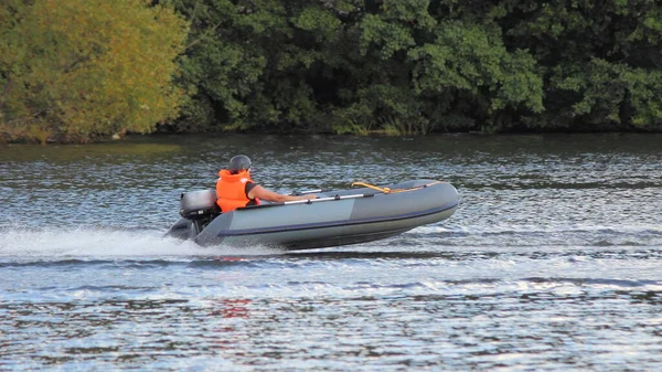 Grey Inflatable outboard motor boat with racer in brighy orange lifejacket and helmet fast glide floats on water Race at river on shore trees background at Sunny summer day, outdoor sport competition — Stock Image Grey Inflatable Outboard Motor Boat Racer Brighy Orange Lifejacket Helmet Stock Picture