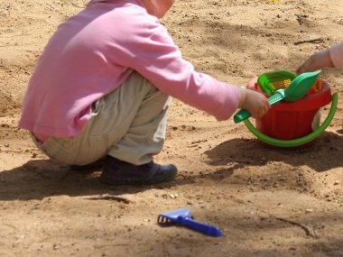 Üç yaşında, açık renk pantolonlu, kumlukta kürek ve kovayla oynayan pembe ceketli, güneşli bir bahar gününde oyun parkında eğlenen Avrupalı bir çocuk.