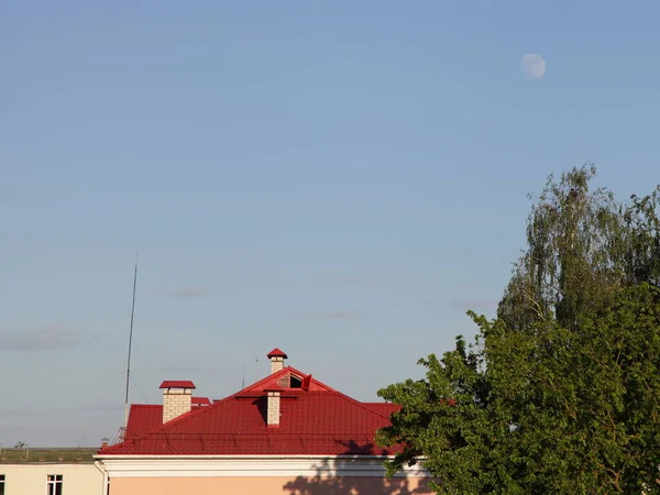Red roof of the house and the green top of the tree with the full moon in the clear blue sky on a summer day